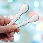 Close-up of a hand holding two white bent utensils resembling spatulas against a blurred background with colorful bokeh lights, reminiscent of the sleek design found in an Aneros Peridise Anal Stimulators (2-Pack) by Aneros.